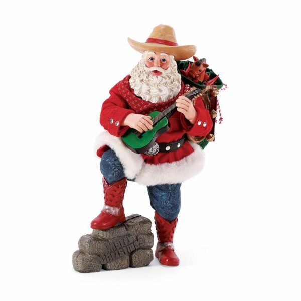 A Cowboy Santa playing a guitar with his pack on his back and his foot on a bale of hay.
