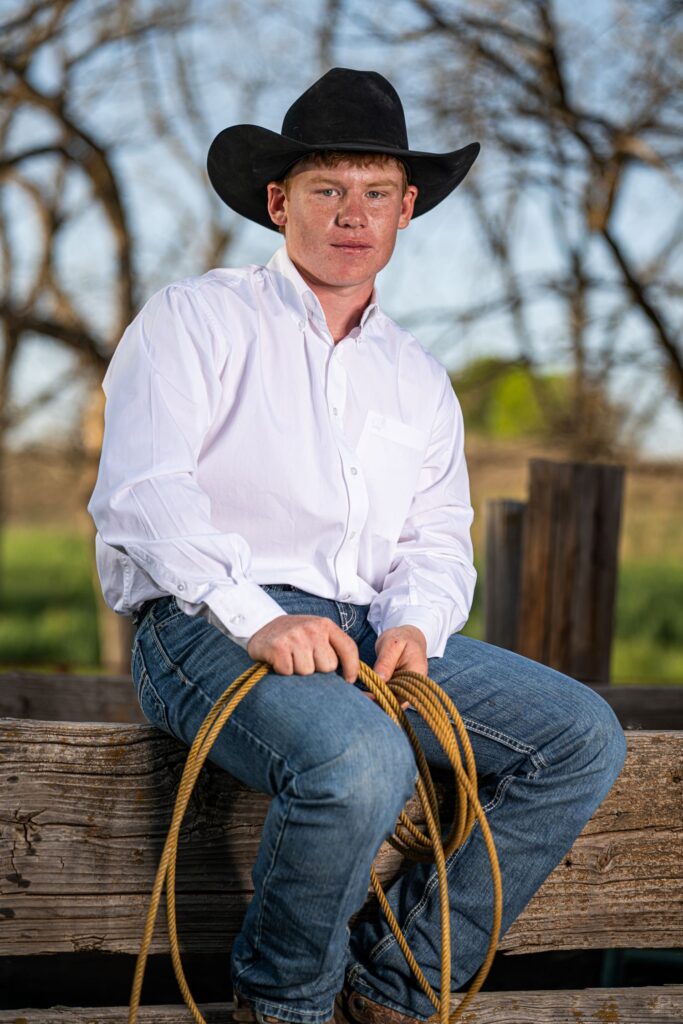 Chizm Kuykendall sits on a board fence wearing a black cowboy hat and holding a rope.